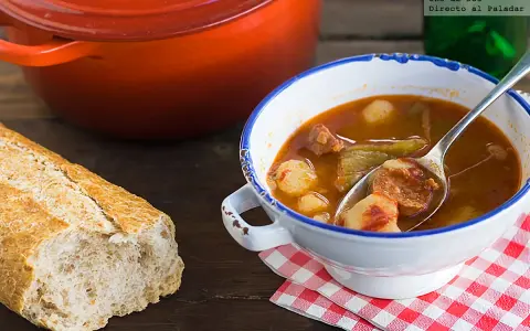 Un reconfortante plato de patatas a la riojana, con chorizo y pimientos, servido en un cuenco blanco con borde azul, junto a una hogaza de pan rústico.