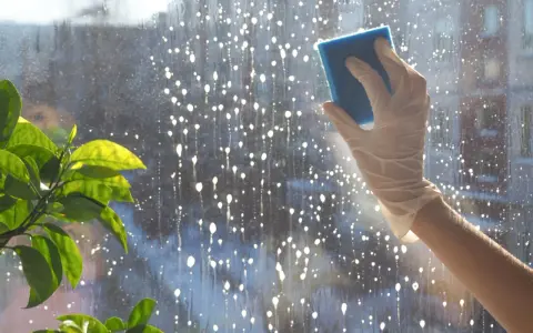 Hand mit Handschuh putzt Fenster mit Schwamm. Tropfen und Blasen auf der Scheibe deuten auf das Ziel hin: Fenster putzen ohne Schlieren.