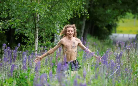 En man med lockigt hår och tatueringar står i en äng full av lila lupiner. Han ser ut att njuta av naturen, kanske som en del av ett avsnitt av "spring snyggt podden".