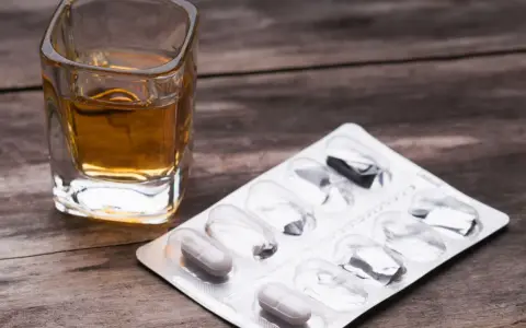 Ein Glas mit bernsteinfarbener Flüssigkeit und eine Blisterpackung mit einigen verbleibenden Pillen. Die Kombination von paracetamol und alkohol kann gefährlich sein.