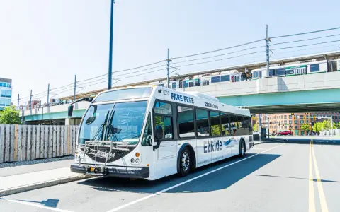 A white EZRide bus, offering fare-free transit from Boston to Cambridge, MA, drives past an elevated train station.