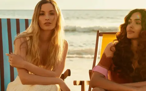 Zwei Frauen entspannen sich am Strand. Die eine mit blonden Haaren, die andere mit braunen Locken. Perfekt für die Haarpflege im Sommer.
