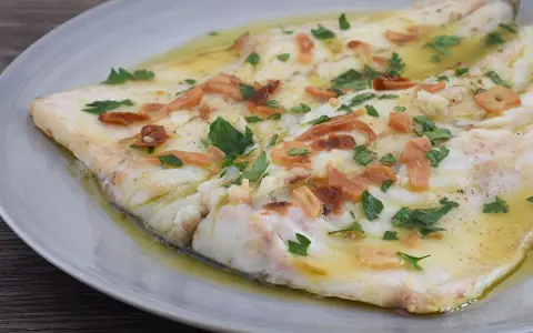 Pescado blanco cocinado en aceite con ajo laminado y perejil picado, servido en plato gris.