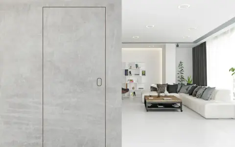 Minimalistyczne wnętrze z betonowymi ścianami. Widoczne drzwi ukryte z niewidoczną klamką, prowadzące do salonu z białą kanapą i stolikiem kawowym.