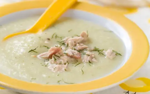 Un cremoso puré de verduras con pollo desmenuzado y eneldo, servido en un plato amarillo con una cuchara.