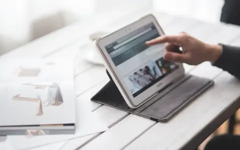 Dłoń wskazuje na ekran tabletu, który wyświetla stronę internetową. Obok leży otwarta książka z grafikami. W tle widać filiżankę. To idealne miejsce do pracy nad projektem, gdzie każdy element, nawet **padding CSS**, ma znaczenie.
