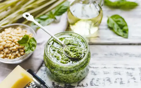 Un barattolo pieno di profumato pesto alla genovese, con pinoli, basilico e olio d'oliva pronti per essere mescolati.