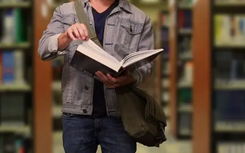 Student w dżinsowej kurtce przegląda książkę w bibliotece. W tle widać regały z książkami.