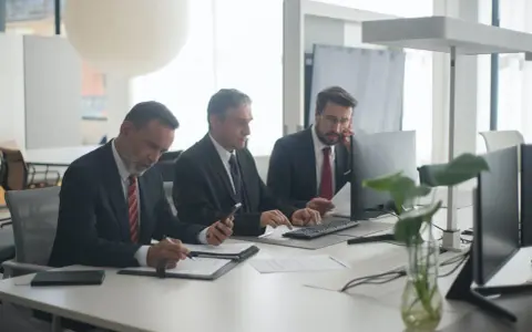 Trzech mężczyzn w garniturach przy biurku. Jeden pisze, drugi patrzy na telefon, trzeci rozmawia przez telefon. Czyżby to byli best s.a. oszuści?