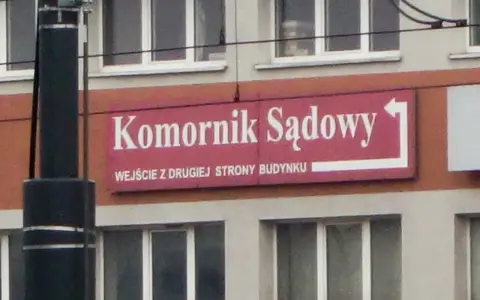 Tablica "Komornik Sądowy" na budynku. Wskazuje wejście z drugiej strony.