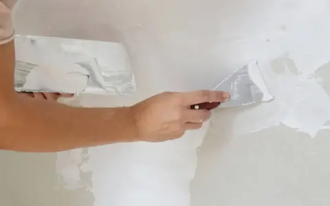 Persona aplicando masilla en una pared con dos espátulas. Aprende cómo masillar una pared para un acabado perfecto.