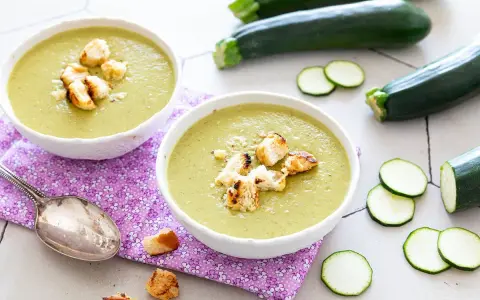 Deux bols de velouté de courgettes crémeux, garnis de croûtons dorés, avec des courgettes fraîches et des rondelles en arrière-plan.
