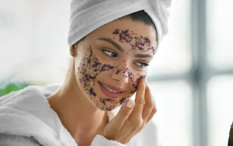 Une femme applique un gommage sur son visage, se demandant combien de gommage par semaine est idéal pour une peau éclatante. Elle porte une serviette sur la tête et un peignoir.