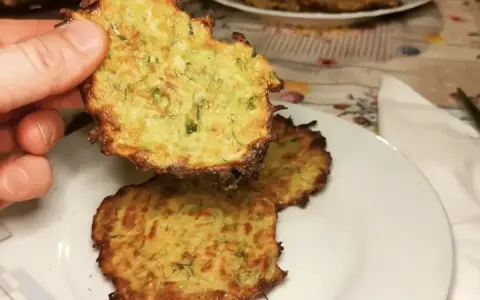 Una mano tiene una frittella di zucchine dorata e croccante. Sul piatto, altre frittelle di zucchine invitano ad essere gustate.