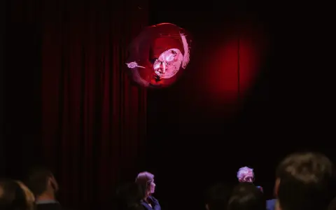 Na scenie, wśród czerwonych zasłon, unosi się kula z projekcją twarzy, przypominająca kurkę wodną. Widownia wpatruje się w to widowisko.