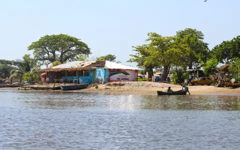 Nad rzeką w Gambii, przy brzegu z łodziami i budynkami, widać tablicę "Welcome to THE GAMBIA".