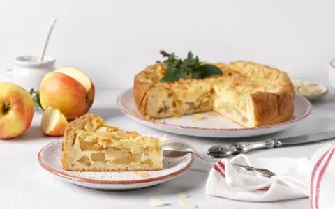 Un délicieux gâteau aux pommes, garni d'amandes effilées, est présenté avec une part déjà coupée. Des pommes fraîches sont disposées à côté.