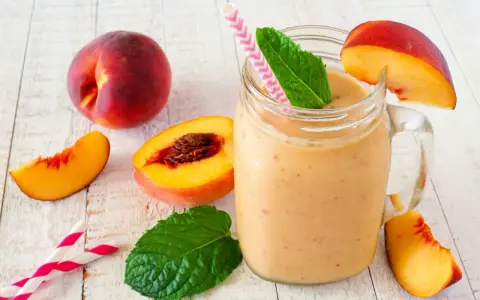 Erfrischender Pfirsich Smoothie im Glas, garniert mit Minze und Pfirsichspalten. Dazu frische Pfirsiche und Trinkhalme.