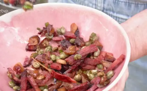 Un plato rosa lleno de habas con jamón, guisantes y un chile rojo seco.