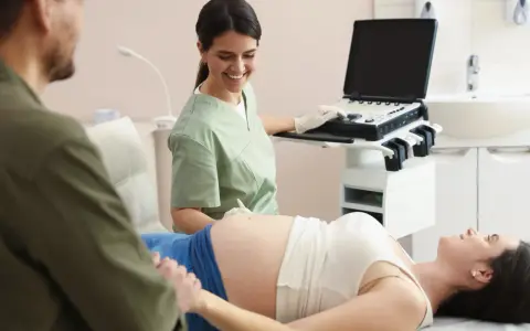 Uśmiechnięta lekarka wykonuje badanie wolnego płodowego dna u ciężarnej pacjentki, podczas gdy partner wspiera ją, trzymając za rękę.