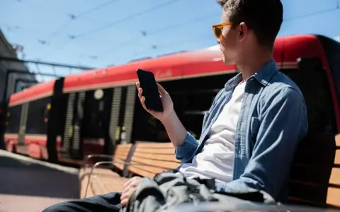 Mężczyzna w okularach siedzi na ławce i trzyma telefon. W tle czerwony tramwaj.