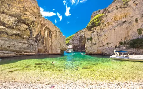 Spiaggia di ciottoli e acque cristalline tra le rocce dell'isola di Lissa, con persone che nuotano e barche.