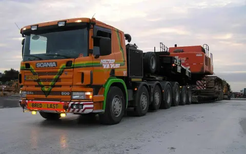 Pomarańczowy Scania z naczepą przewozi koparkę. Transport ponadgabarytowy wymaga specjalnych godzin jazdy.