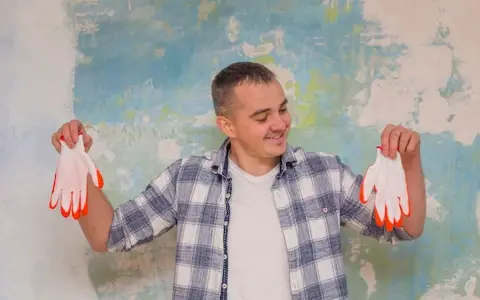 Homme souriant tenant des gants blancs à bouts orange devant un mur texturé. Il semble prêt à peindre, peut-être pour estimer le prix peinture m2 au black.