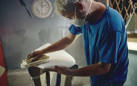 Artisan masqué ponçant une planche de surf blanche, étape cruciale avant de faire de l'époxy.