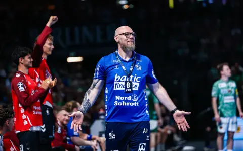 Trener SG Flensburg-Handewitt w niebieskiej koszulce z tatuażem na ramieniu, z otwartymi dłońmi, podczas meczu.