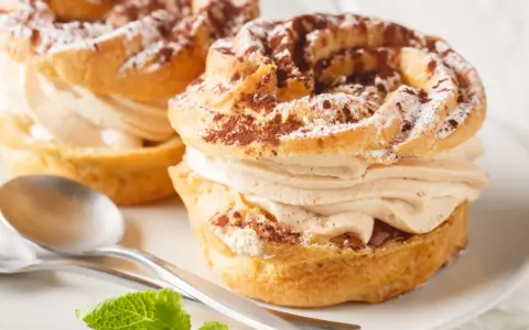 Dwa pyszne ciastka Paris-Brest z kremem kawowym, posypane kakao i cukrem pudrem, na talerzu ze srebrnymi łyżeczkami i miętą.