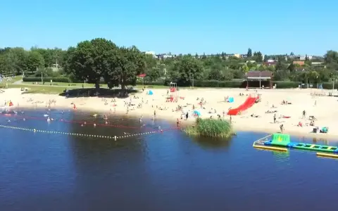 Słoneczny dzień nad jeziorem Grzymisławskim. Ludzie odpoczywają na plaży, kąpią się i bawią na dmuchanych atrakcjach.