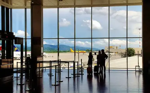 Widok z okna na lotnisko Bergamo. Trzy osoby z walizkami czekają na samolot. W tle żółty samolot DHL i góry.