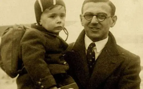 Nick Winton porte un enfant sur son bras, un petit garçon avec un sac à dos, prêt pour un nouveau départ.