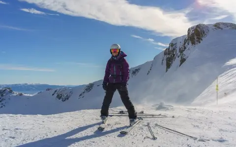 Narciarka w fioletowej kurtce i goglach stoi na ośnieżonym szczycie góry.