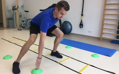 Mężczyzna w niebieskiej koszulce ćwiczy zwinność na drabince koordynacyjnej. 4 miesiące po rekonstrukcji ACL, wraca do formy.