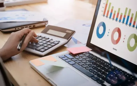 Main d'une personne utilisant une calculatrice pour le budget projet informatique, avec un ordinateur affichant des graphiques.