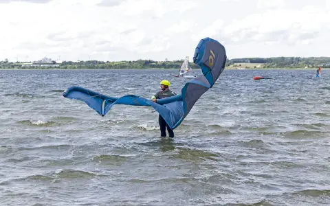 Osoba w żółtym kasku przygotowuje się do **kite surfingu**, trzymając duży, niebiesko-szary latawiec na wzburzonej wodzie.