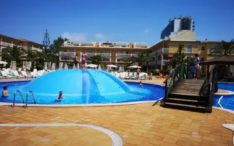 Sole e divertimento in piscina a Maiorca con bambini. Una grande struttura gonfiabile blu al centro, bagnanti e hotel sullo sfondo.