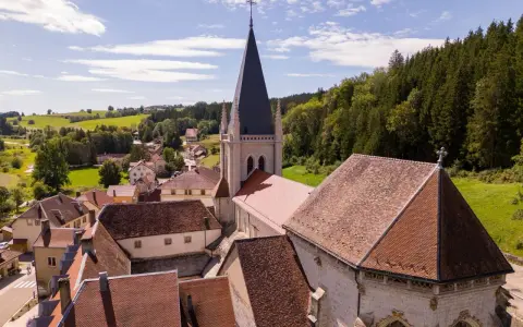 Vue aérienne d'un village pittoresque du haut Doubs, dominé par une église à la flèche élancée, entouré de collines verdoyantes et de forêts.