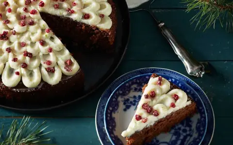 En mjuk pepparkaka med fluffig frosting och röda bär. En bit är upplagd på en blåvit tallrik, redo att avnjutas.