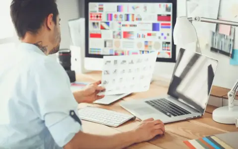 UX UI designer pracuje przy biurku, analizując wydruki i ekran komputera z paletą barw.