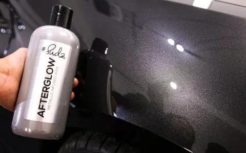Bottle of "Afterglow Metallic Infused Wax" held against a dark, metallic car finish, showcasing the product's ability to create multiple coats of wax for a deep shine.