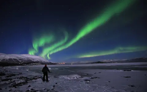 En ensam person står på en isig strand och ser på det magiska abisko norrsken som dansar över himlen.