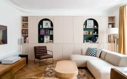 Un salon moderne avec un canapé beige, une chaise en cuir marron et des étagères murales roses. Quelle couleur pour une grande pièce à vivre ? Le vert émeraude des niches apporte une touche audacieuse.