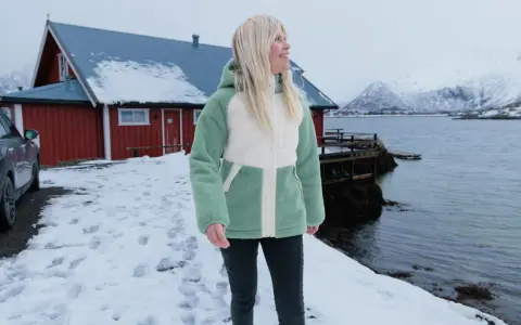 Frau in flauschiger Fleecejacke, bereit für das **fleece waschen**, steht vor rotem Haus am Meer.
