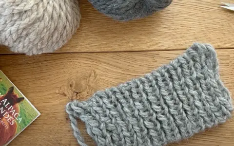 Ovillos de lana gris y beige sobre madera. Un trozo de tejido con puntos de lana muestra la textura.