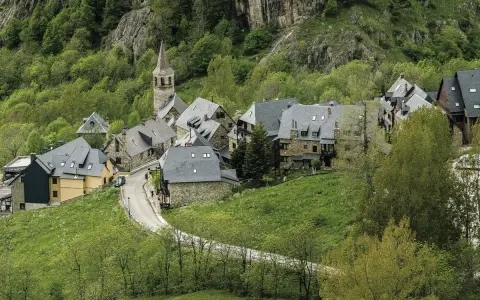 Pueblos del Valle de Arán, con casas de piedra y tejados grises, se asoman entre la vegetación verde y un camino serpenteante.