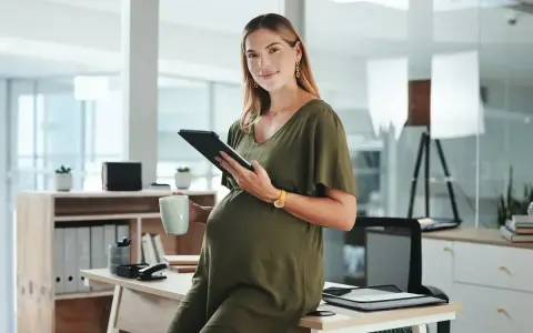 Uśmiechnięta ciężarna kobieta z tabletem i kubkiem w biurze. Dowiedziałam się o ciąży po okresie wypowiedzenia, ale teraz planuję przyszłość.