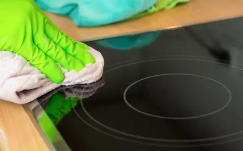 Grüne Handschuhe wischen mit einem Tuch über das Induktionskochfeld, um es zu reinigen.
